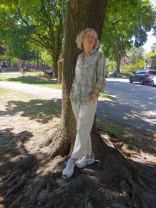 A photo of a woman leaning against a tree. She is wearing a light coloured checked shirt and white pants. She has white wavy har and glasses.